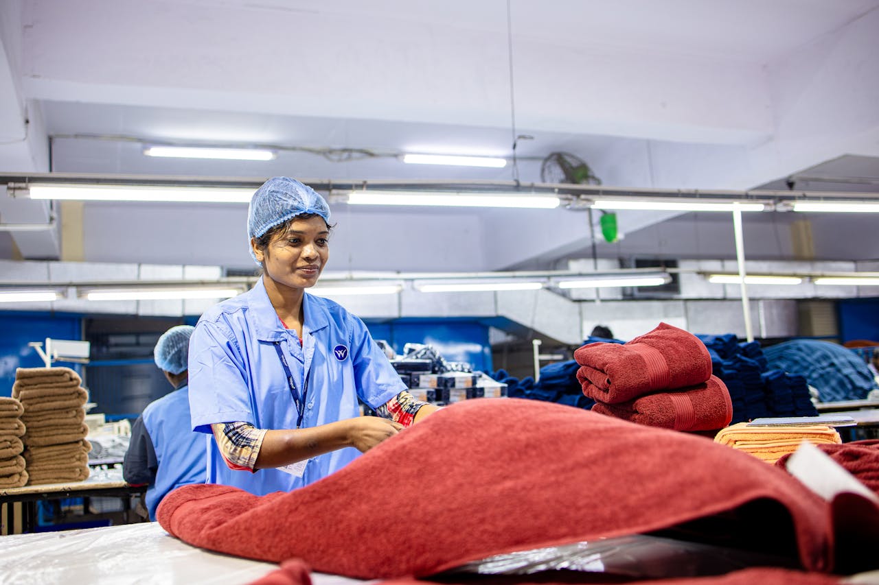 Smiling female factory worker folding towels in a busy textile manufacturing plant.